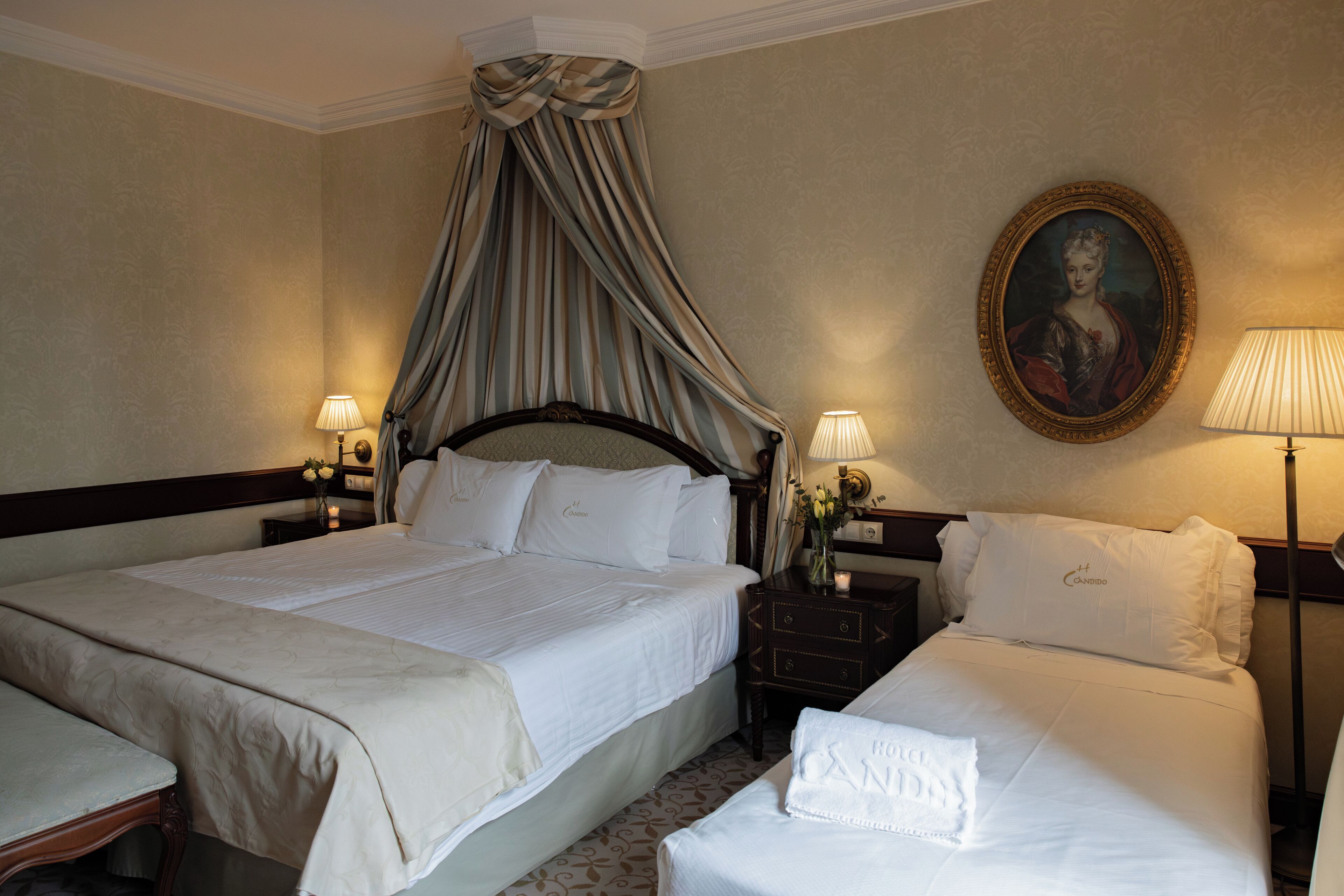A hotel room with two beds, a canopy bed, a nightstand with a lamp, a framed portrait, and a vase with flowers.