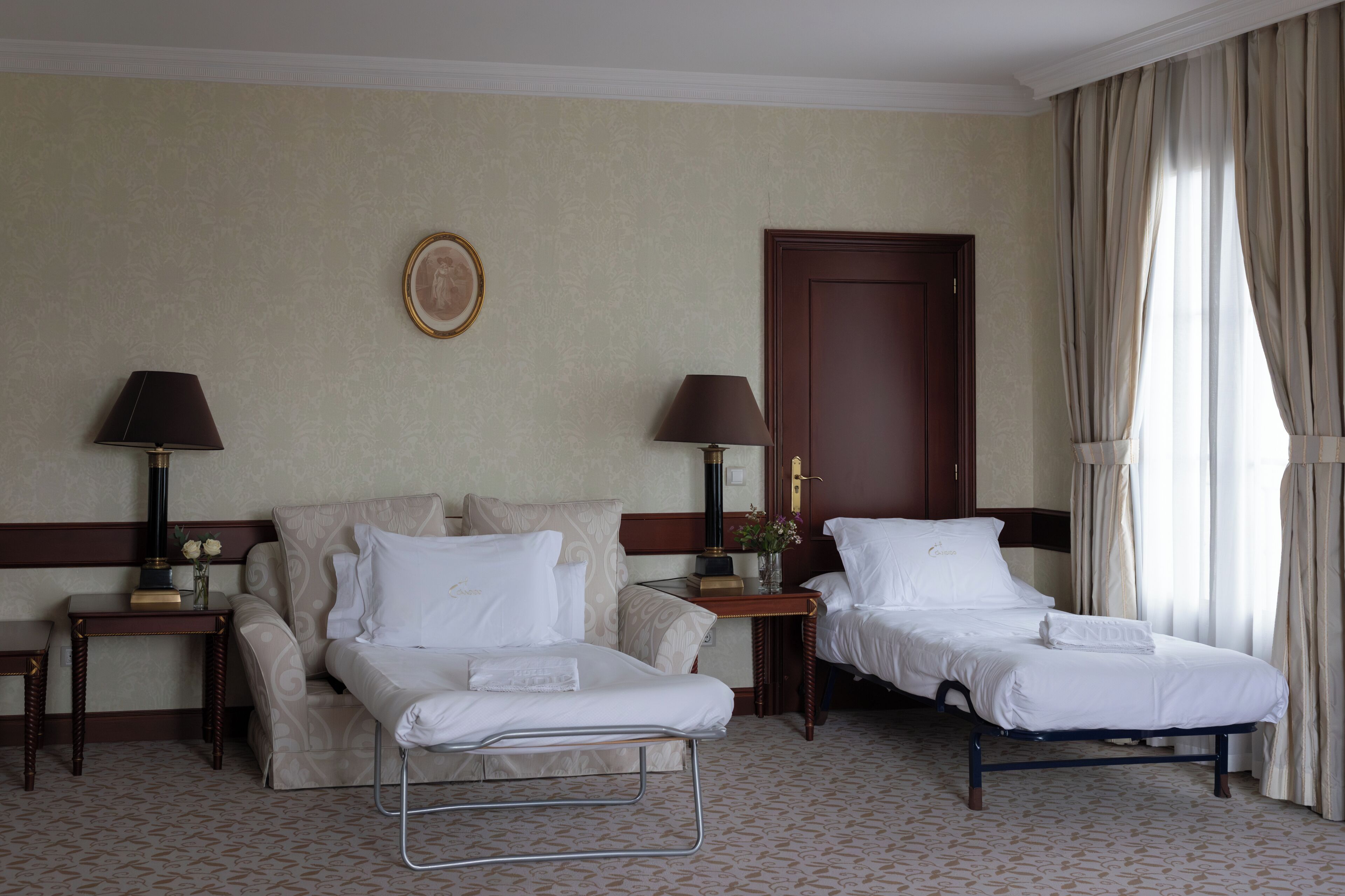 A hotel room with two beds, a bedside table with a lamp, a wall-mounted picture, and a door with curtains.