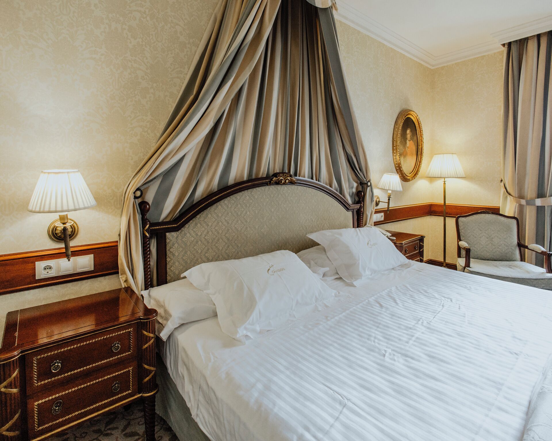 A hotel room with a four-poster bed, a nightstand with a lamp, a chair, and a mirror on the wall.