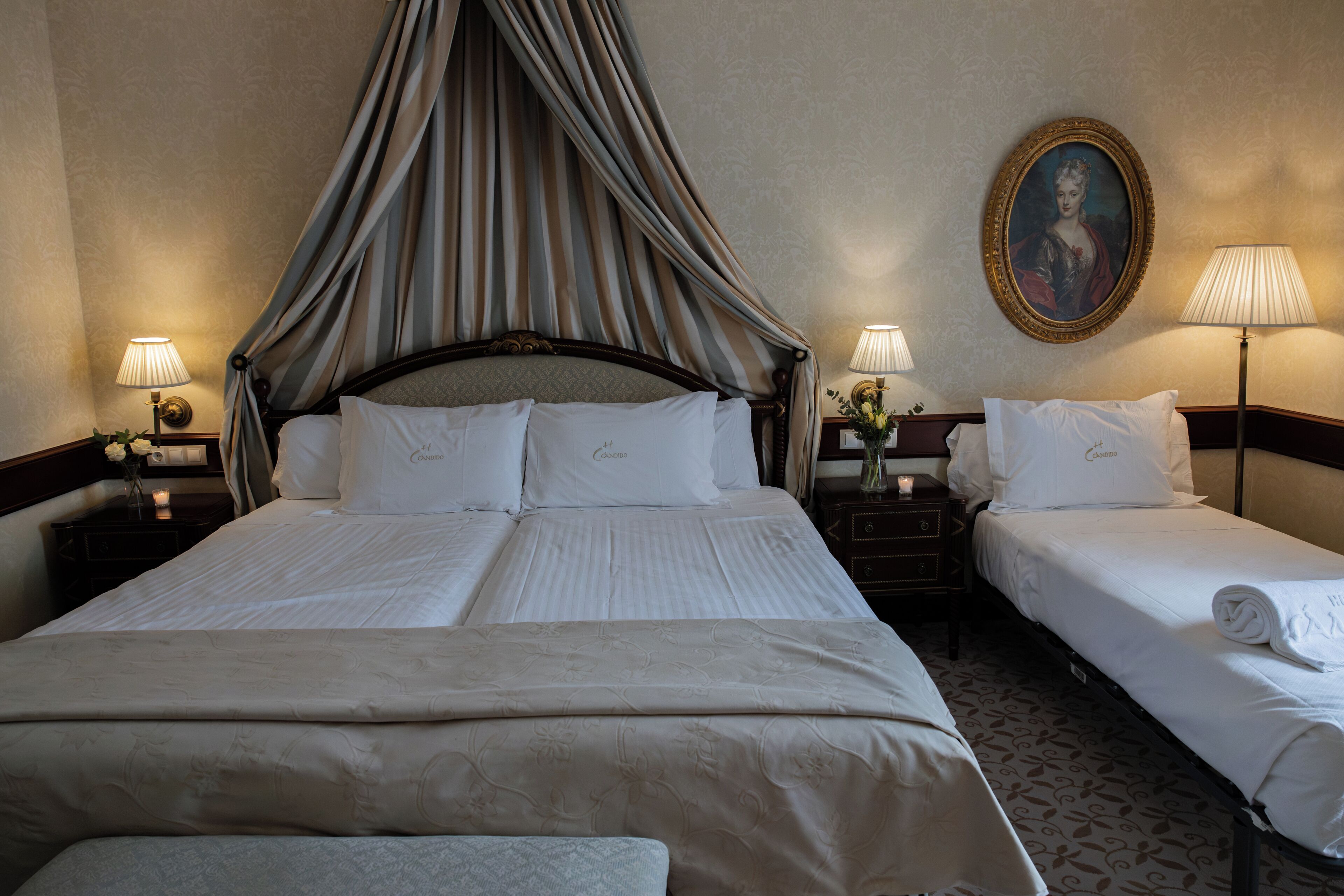 Een hotelkamer met een hemelbed, twee nachtkastjes, een lamp, een bloemenvaas en een lijst met een portret aan de muur.