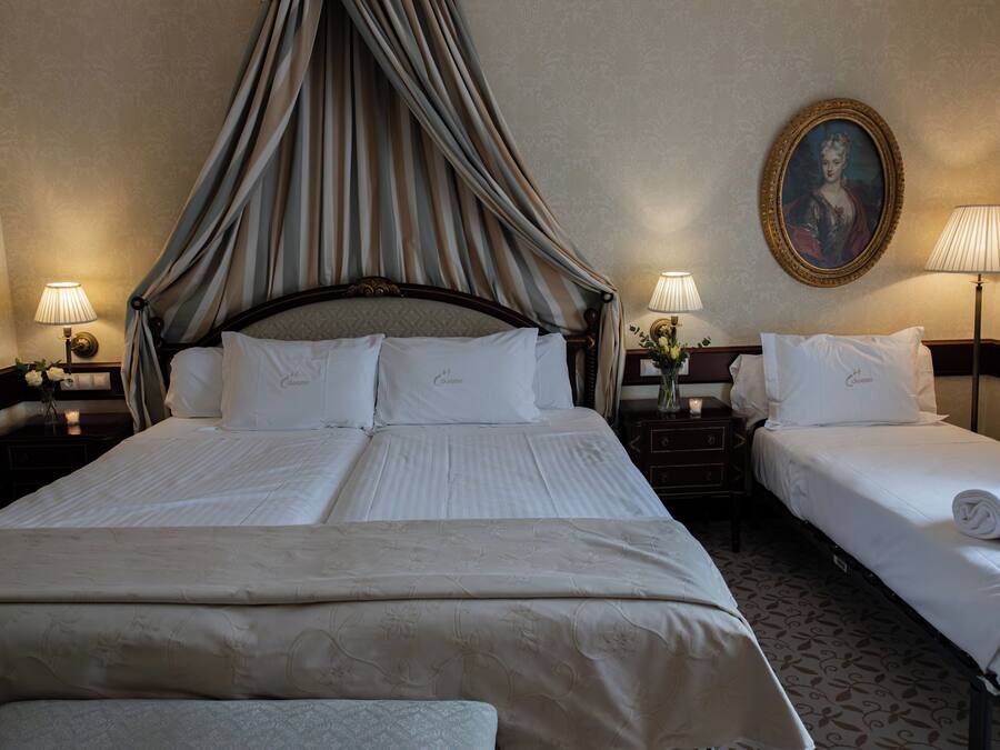 A hotel room with a four-poster bed, two bedside tables, a nightstand, a lamp, a vase with flowers, and a framed portrait on the wall.