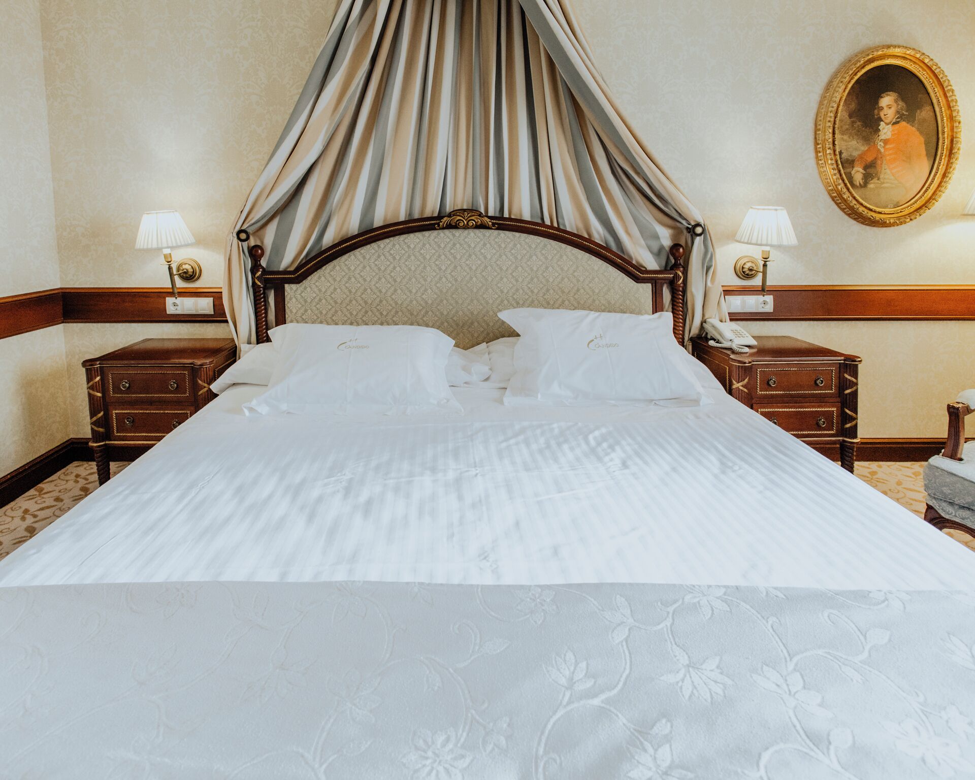 A four-poster bed with a canopy, two bedside tables with lamps, a nightstand with a telephone, and a framed portrait on the wall.