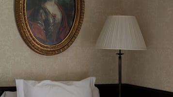 Una cama bien hecha con sábanas blancas y una toalla enrollada, un retrato enmarcado en la pared y una lámpara de pie junto a la cama.