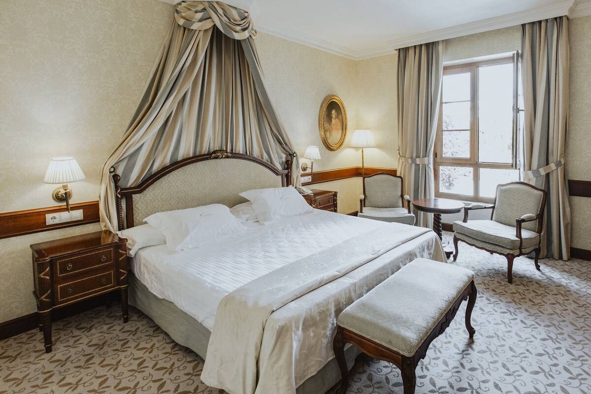 A hotel room with a large bed, a bedside table, a chair, a small table, and a window with curtains.
