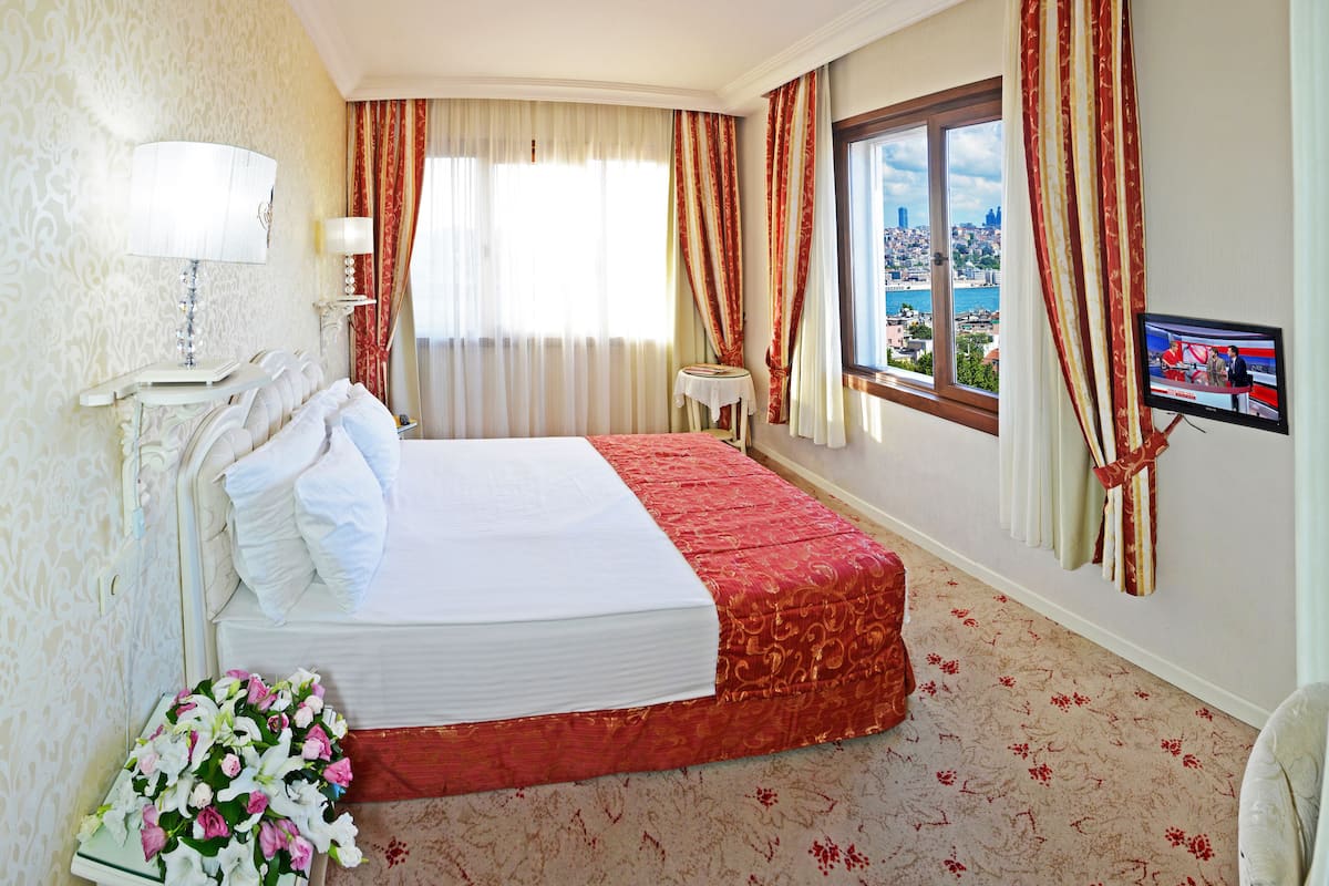 A hotel room with a bed, a TV, a window with a city view, and a flower arrangement.