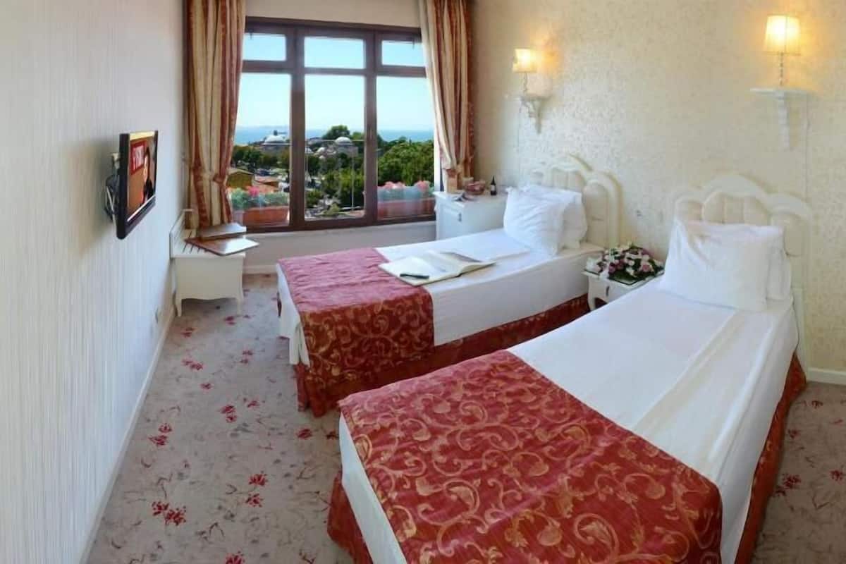 A hotel room with two beds, a window with a view, a bedside table with a lamp, and a wall-mounted picture.