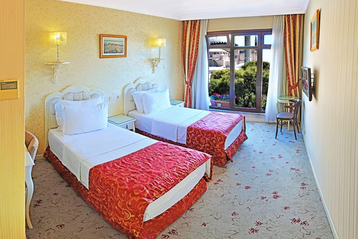 A hotel room with two beds, a small table, a chair, and a window with a view of greenery.