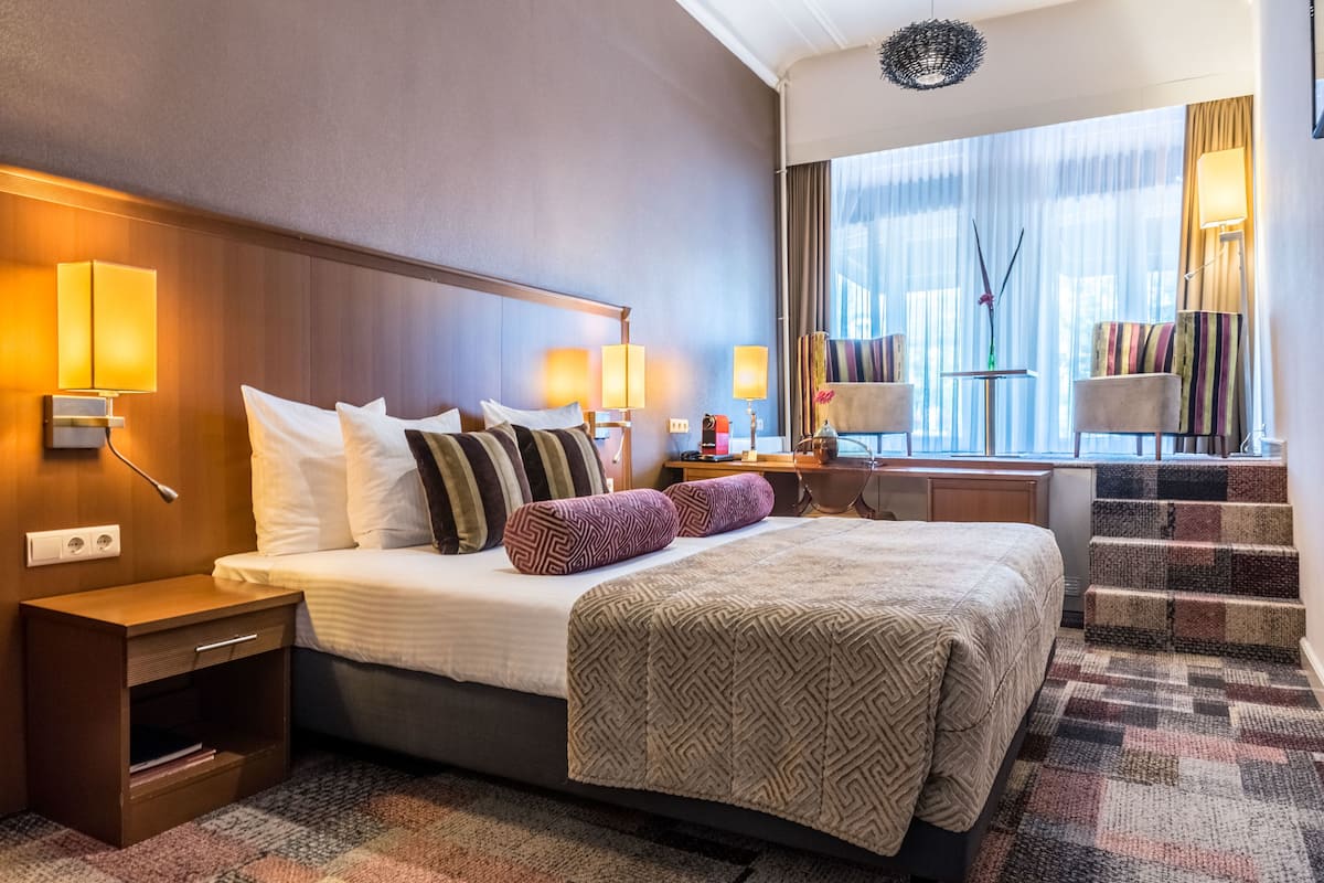 A hotel room with a large bed, two bedside tables, a desk with a chair, and a window with curtains.