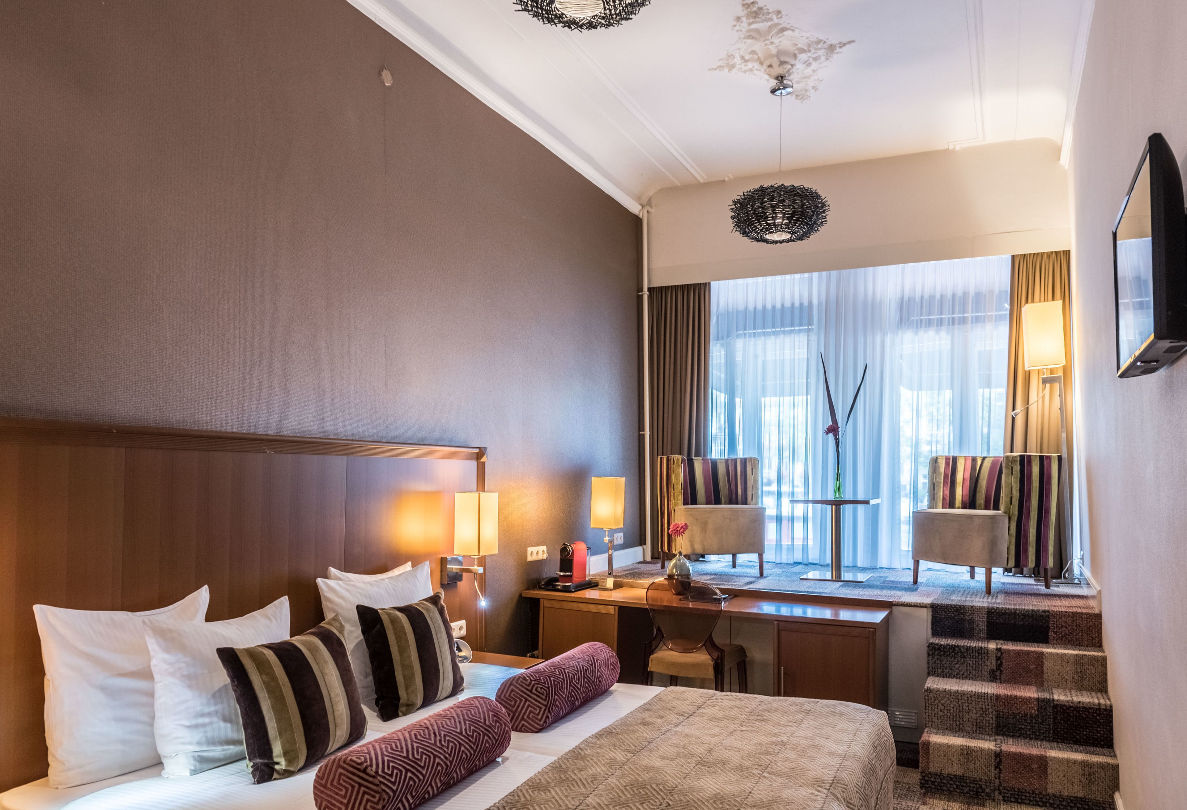 Habitación de hotel con una cama grande, un escritorio, una silla y vistas de la ciudad a través de amplios ventanales.