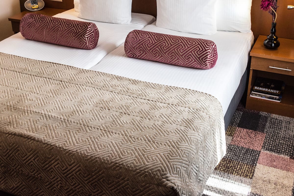 A hotel room with a neatly made bed, a bedside lamp, a nightstand with a vase of flowers, and a small drawer unit.