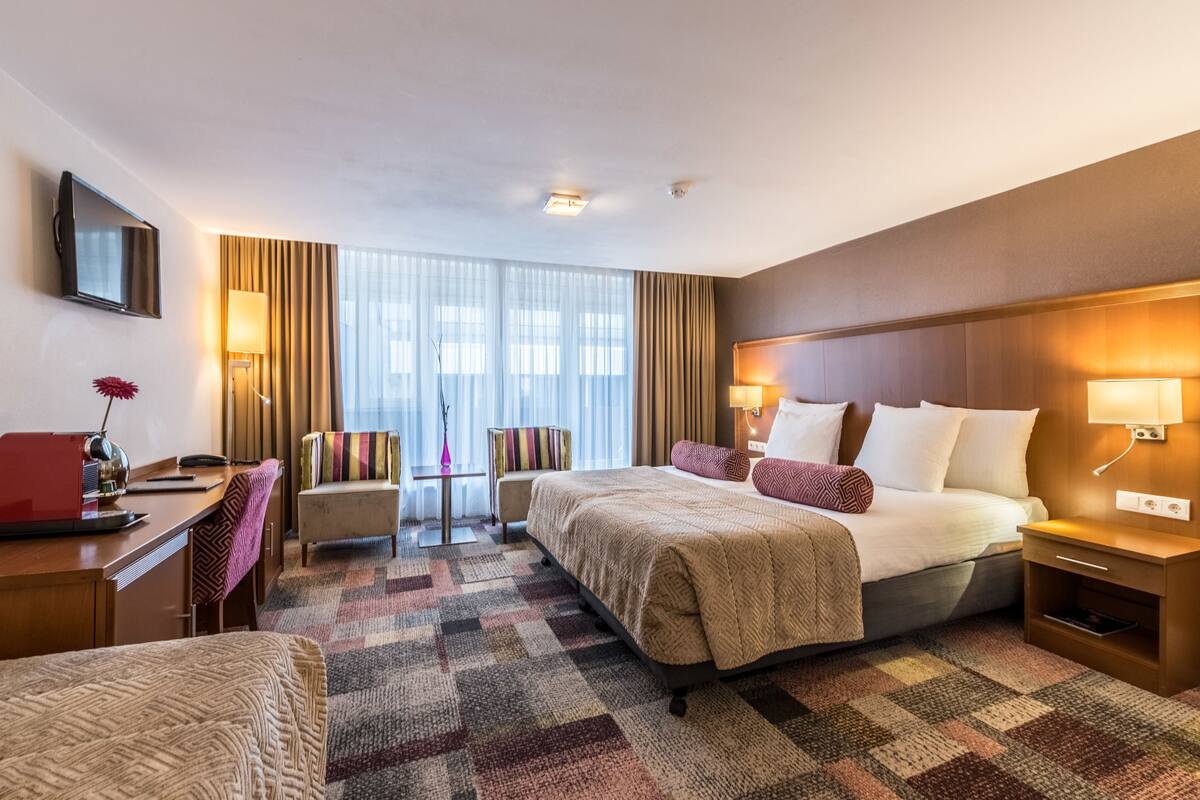 A hotel room with a large bed, a desk, a chair, a TV, and a window with curtains.