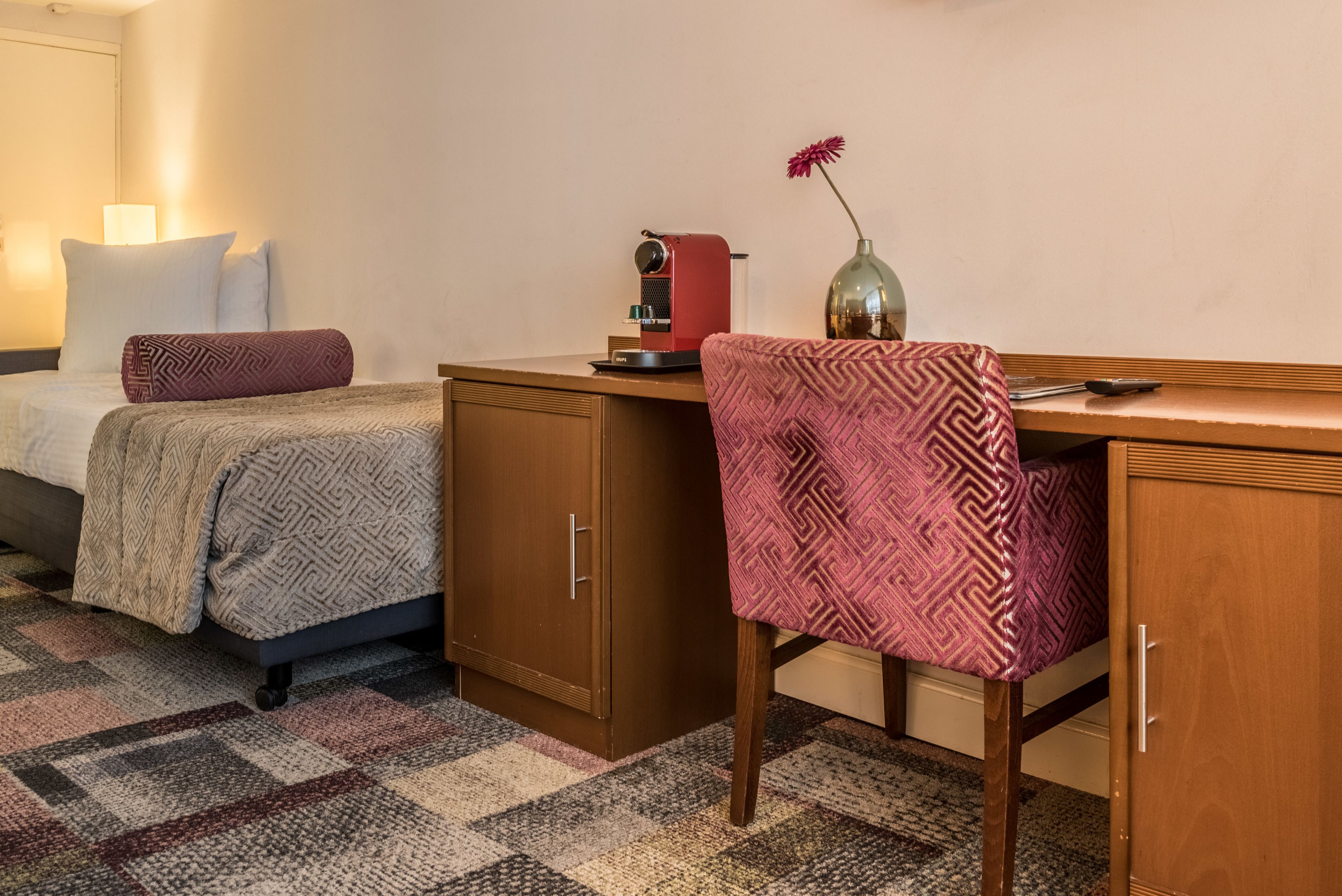 A hotel room with a bed, a desk, a chair, a coffee machine, and a vase with a flower.