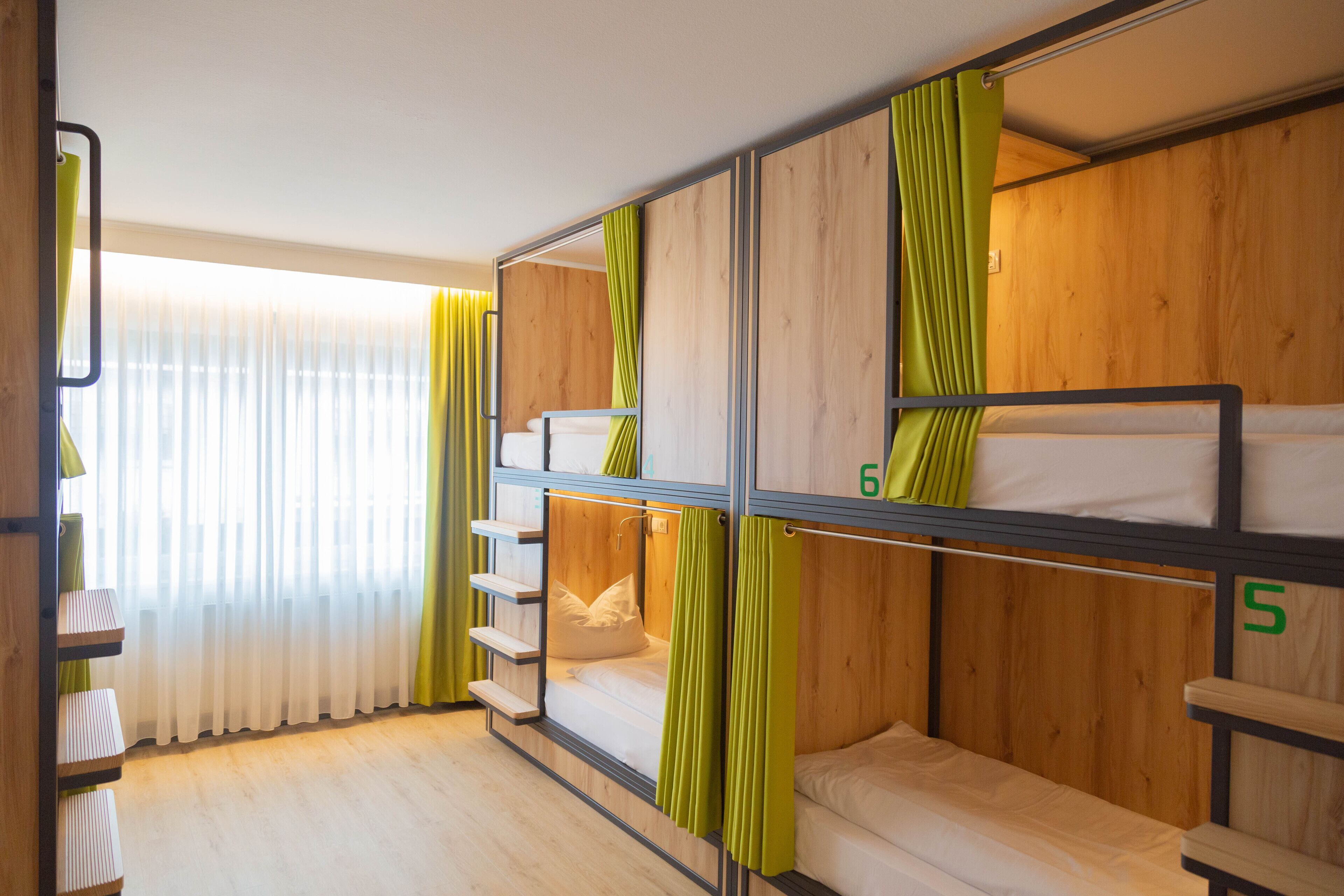 A bunk bed room with wooden walls and green curtains, featuring a window with sheer curtains and a wooden floor.