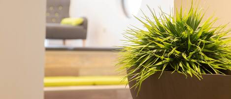 Un interior moderno con una planta en maceta sobre un pedestal, muebles de madera y una vista parcial de una sala de estar al fondo.