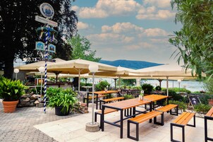 Un café al aire libre con mesas y sombrillas, un letrero que anuncia un jardín de cerveza, y vistas a un cuerpo de agua y montañas.