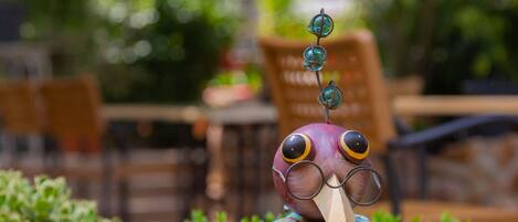 Une décoration en forme d’oiseau, dotée d’un long bec et d’yeux colorés, placée sur un fond flou de verdure et de mobilier d’extérieur.