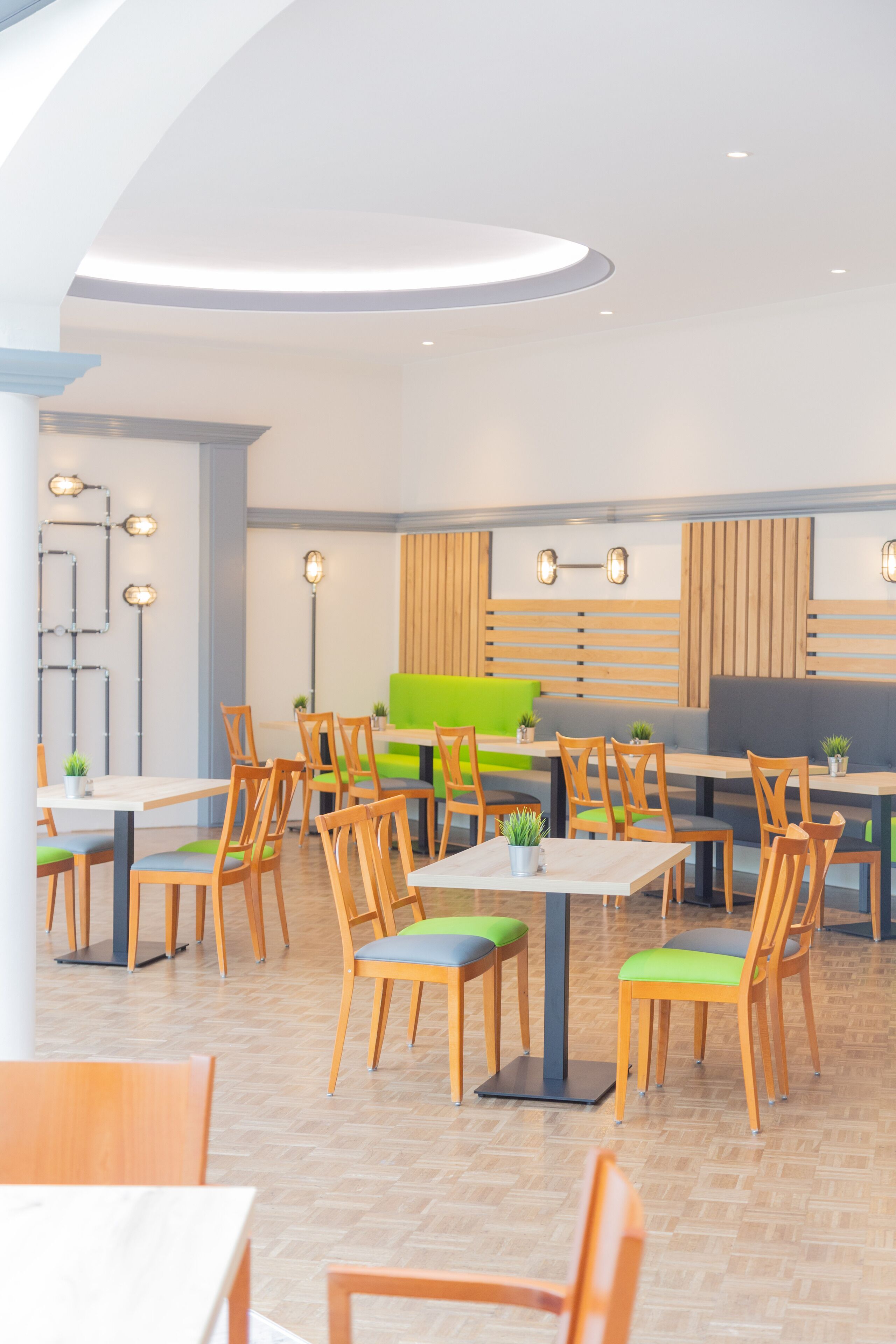 Interior moderno de restaurante con mesas y sillas de madera, asientos acolchados verdes e iluminación colgante.