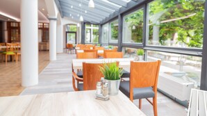A modern restaurant with wooden tables and chairs, a potted plant, salt and pepper shakers, and large windows offering a view of the outdoors. - Das Ebertor Hotel & Hostel (Boppard)