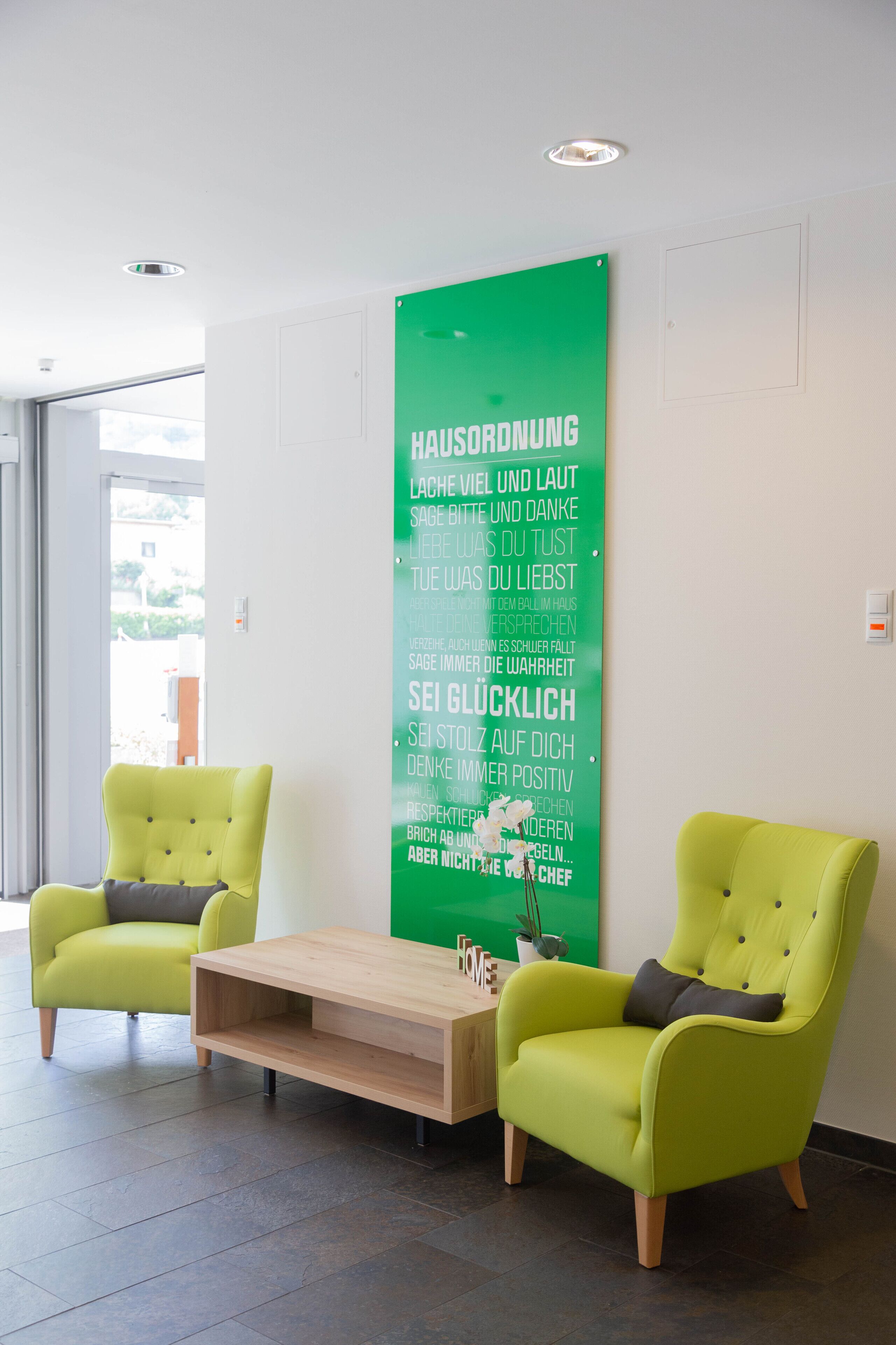 Eine moderne Lobby mit zwei grünen Sesseln, einem Holztisch und einer grünen Wand mit motivierenden Texten.