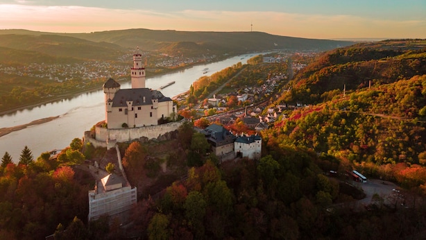 A castle-like building with a tower, surrounded by a forested area and a river. - Das Ebertor Hotel & Hostel (Boppard)