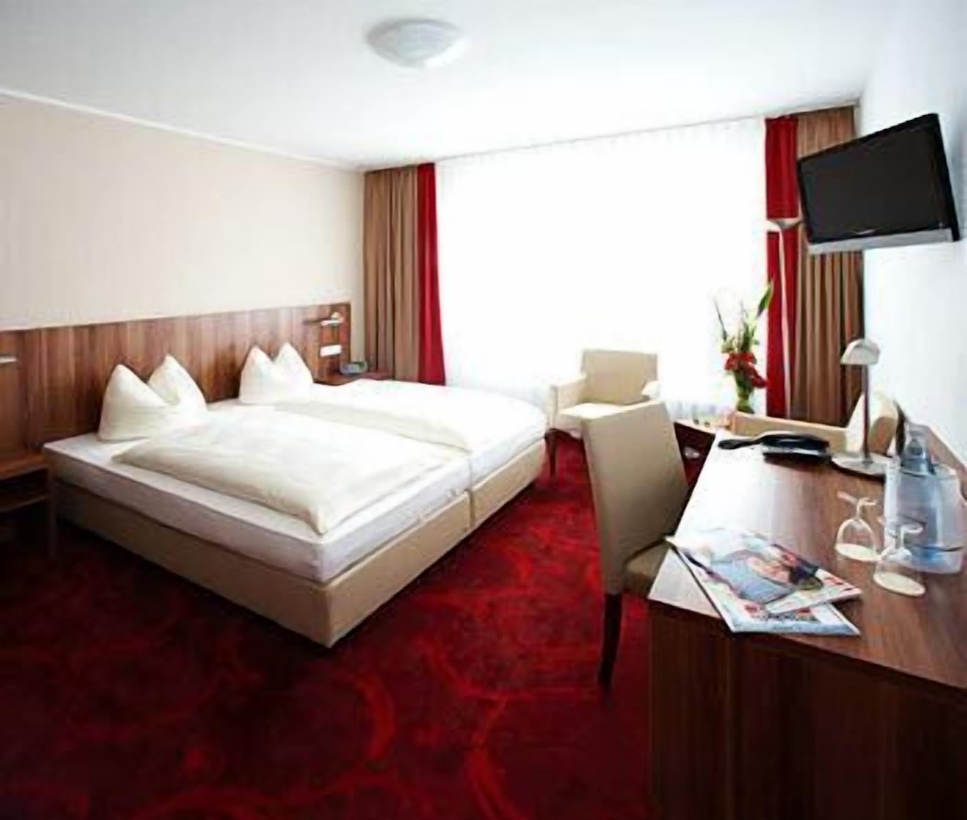 A hotel room with a large bed, a desk, a chair, a TV, and a red carpet.