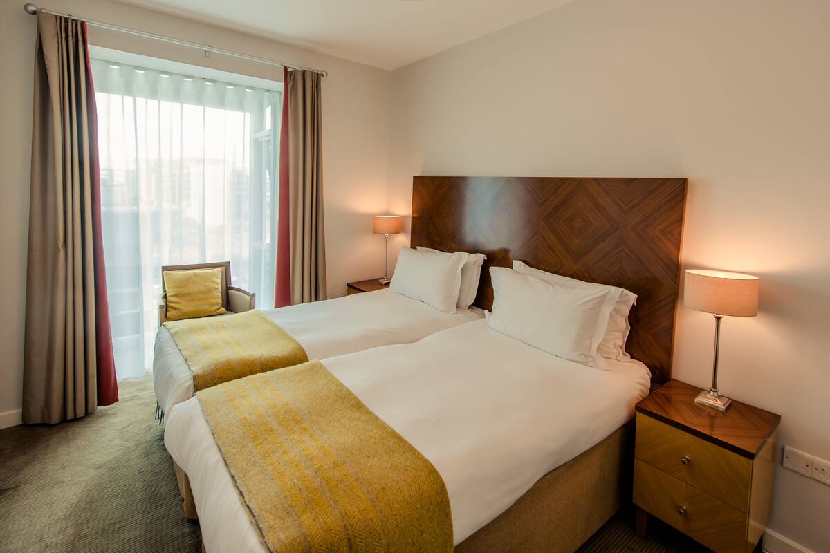 Une chambre d’hôtel avec deux lits, une tête de lit en bois, des lampes de chevet, une chaise et une grande fenêtre avec des rideaux.