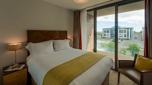 Hotellihuone, jossa on suuri sänky, yöpöytä lampun kanssa, tuoli ja näkymä rakennuksiin ikkunasta.