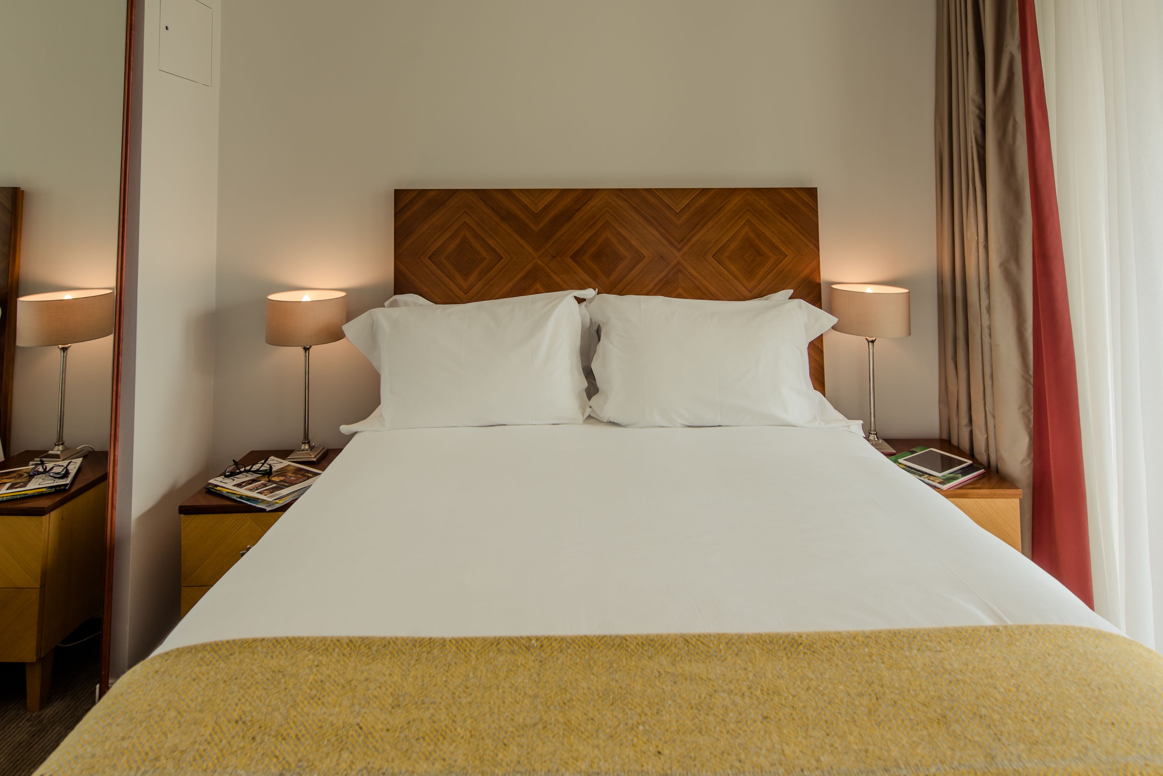 A hotel room with a large bed, two bedside tables, a wooden headboard, and a window with curtains.