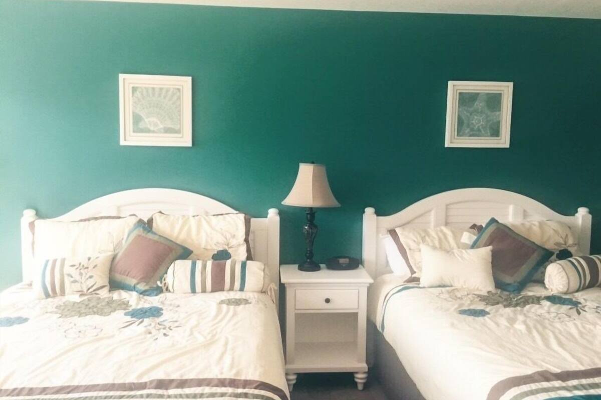 Una habitación con dos camas, cada una con cabecero blanco, una mesita de noche con lámpara y dos cuadros enmarcados sobre la pared color turquesa.