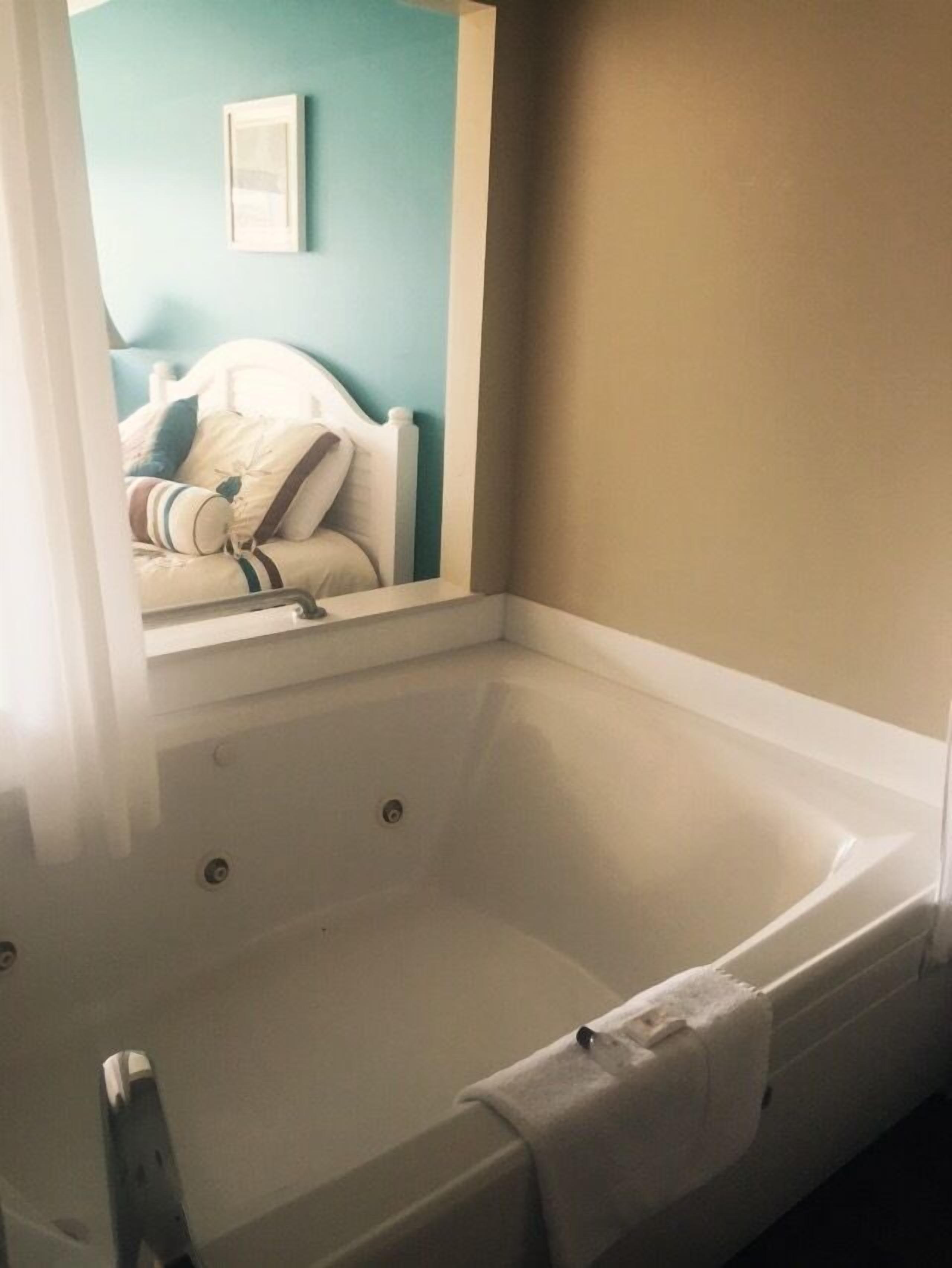 A bathroom with a white bathtub, a mirror reflecting a bed with pillows, and a blue wall.