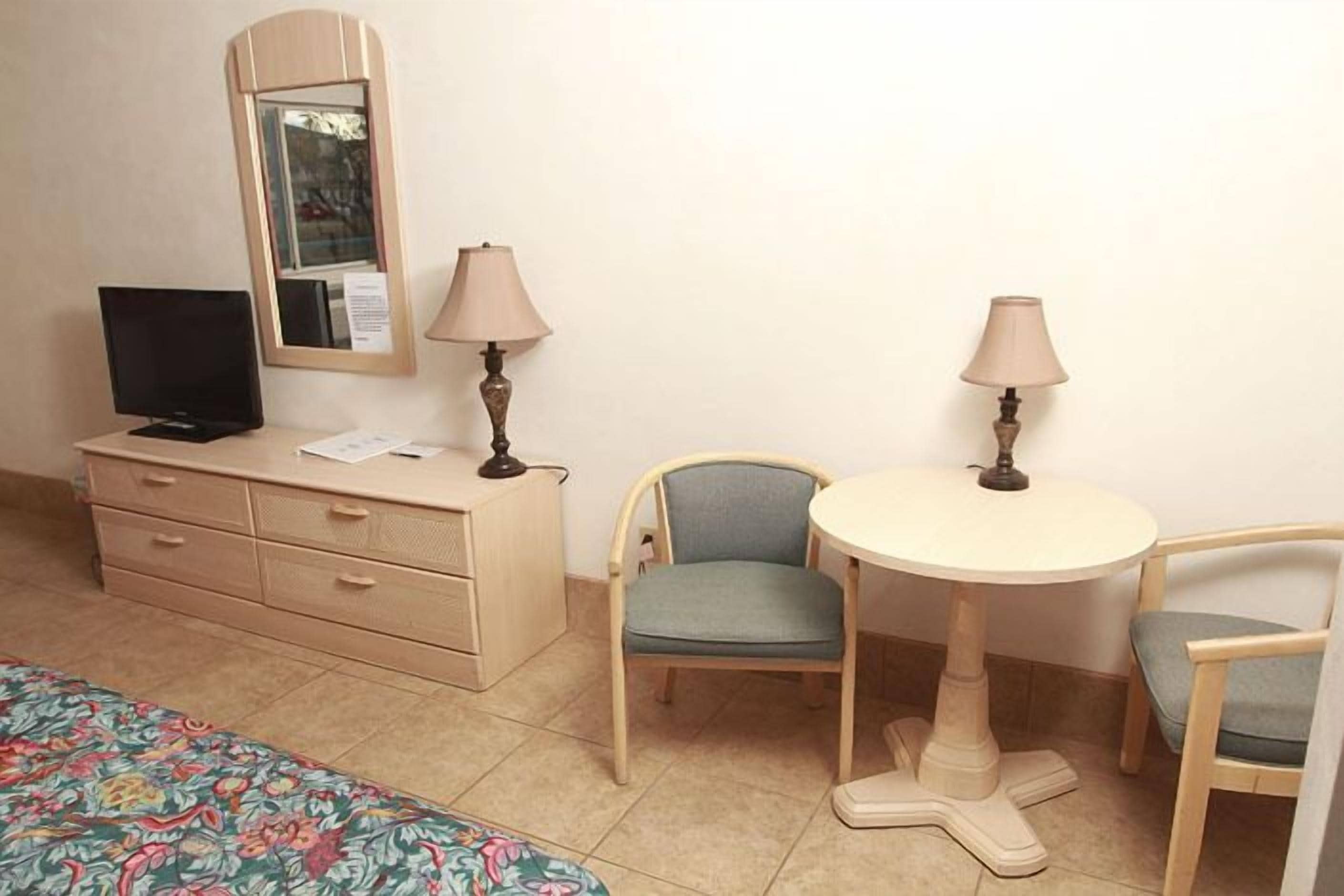A hotel room with a bed, a desk with a computer, two chairs, a small table, and two lamps.