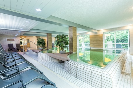 A modern indoor pool area with seating, a water feature, and large windows.. AktiVital Hotel