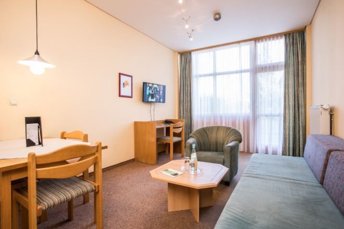 Une chambre d’hôtel avec une table à manger en bois, deux chaises, un canapé, une petite table basse, un téléviseur et une grande fenêtre avec des rideaux.