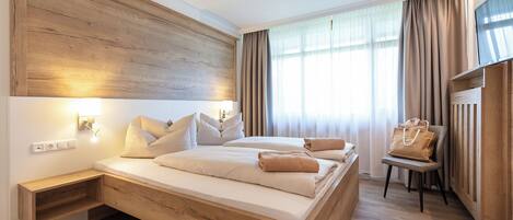 A modern hotel room with a wooden headboard, a large bed with multiple pillows, a bedside table, a chair, and a large window with curtains.