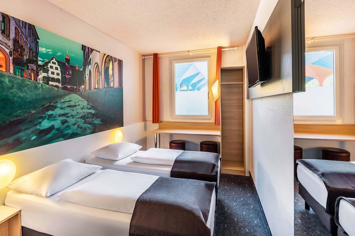 Une chambre d’hôtel avec deux lits, un tableau représentant un paysage urbain, une télévision et une fenêtre avec des rideaux.