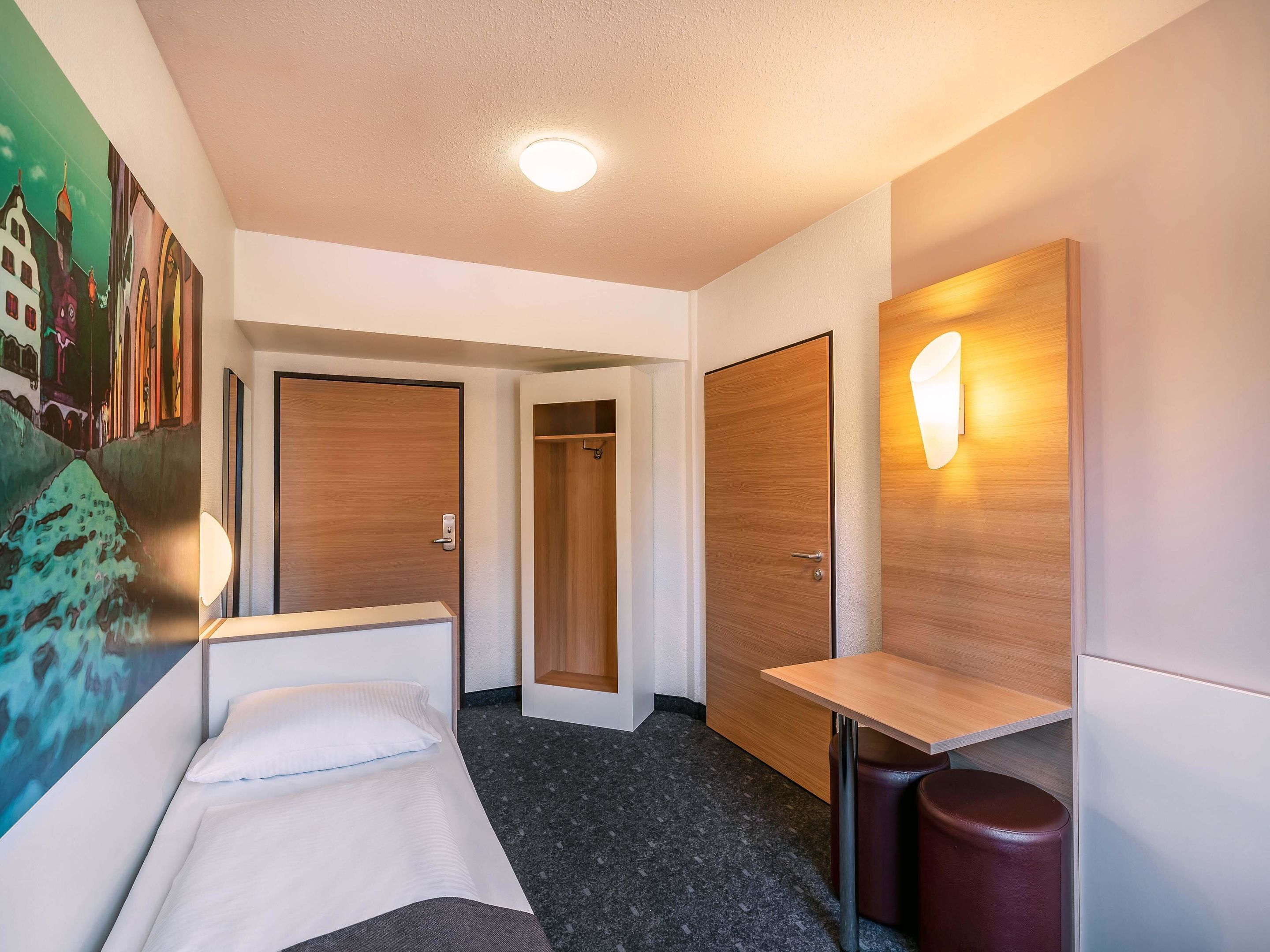 A hotel room with a bed, a desk, a wall-mounted light, and a door.