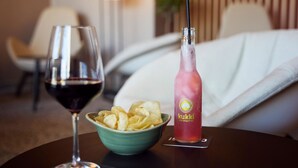 A hotel room with a dining table featuring a glass of red wine, a bowl of chips, and a bottle of pink grapefruit drink. - B&B Hotel Freiburg-Nord (Freiburg im Breisgau)