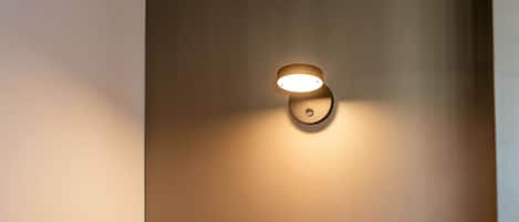 A neatly made bed with a headboard against a plain wall, a wall-mounted light fixture, and a small circular object on the wall.