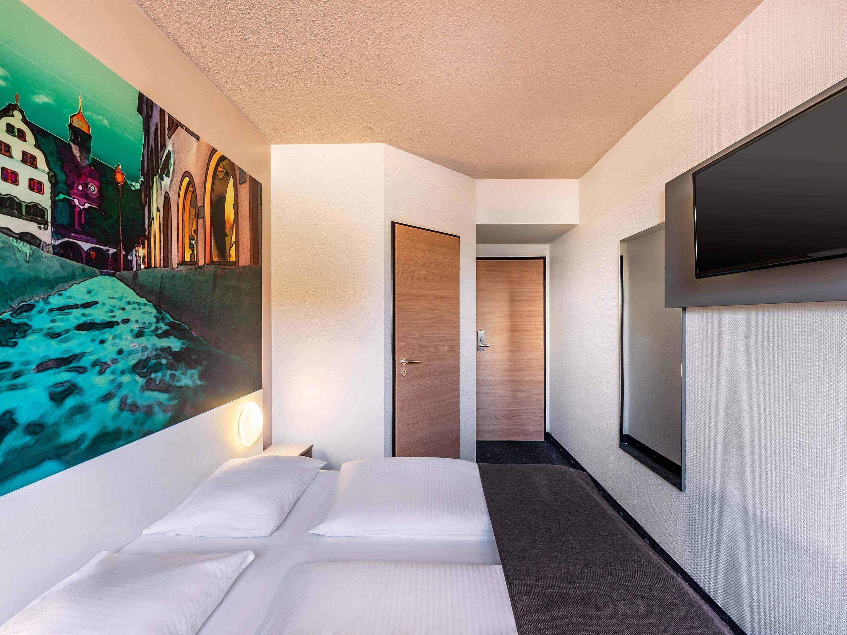 A hotel room with a bed, a wall-mounted TV, and a door leading to another room.