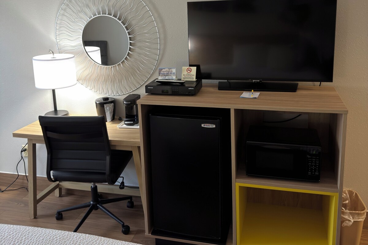A hotel room with a desk, a television, a refrigerator, a microwave, a coffee maker, a trash bin, and a chair.