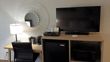 Una camera d'albergo con una scrivania, televisione, frigorifero, forno a microonde, macchina del caffè, cestino e una sedia.
