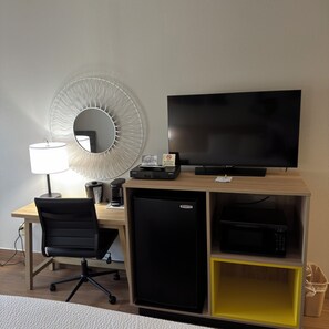 A hotel room with a desk, a television, a refrigerator, a microwave, a coffee maker, a trash bin, and a chair.