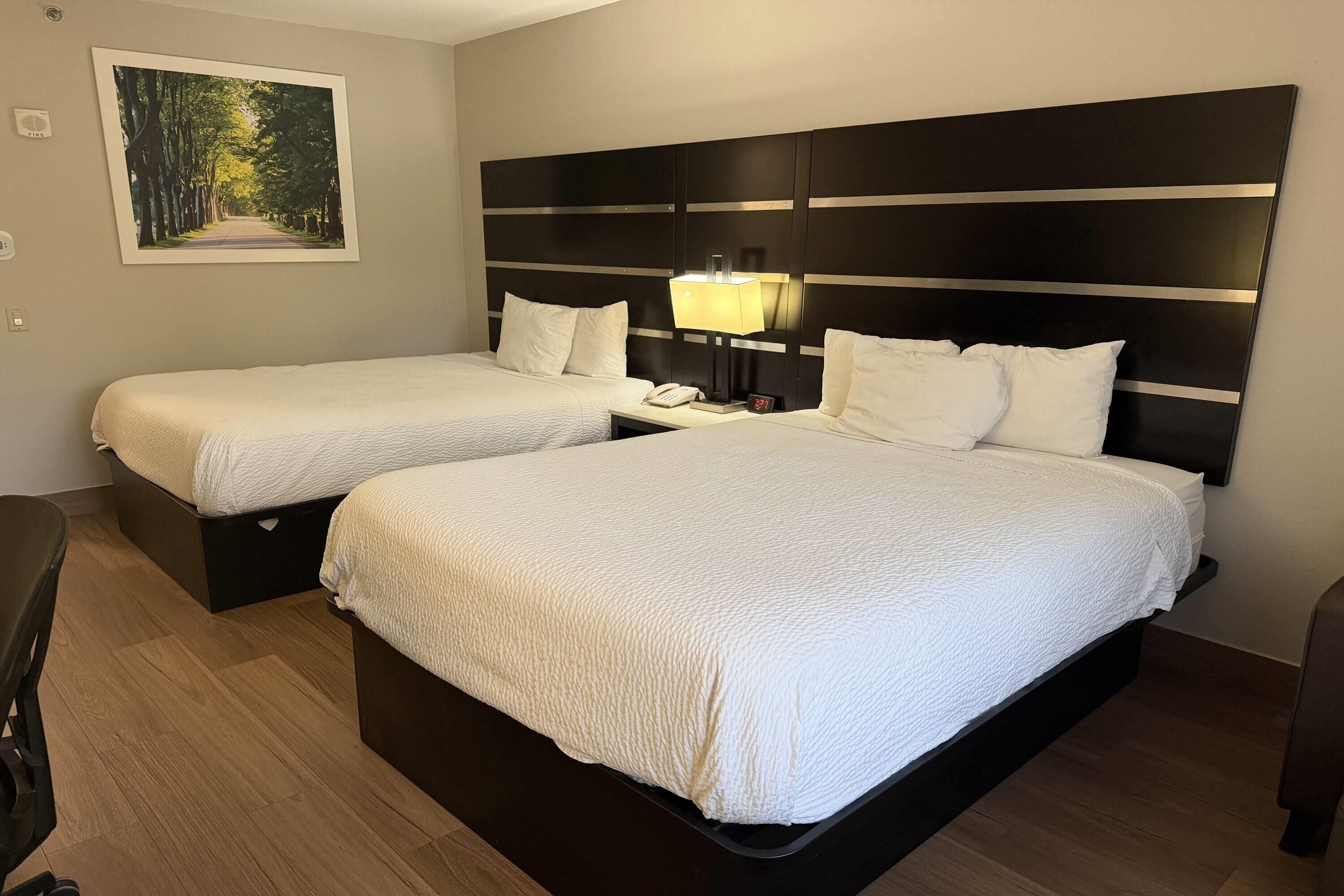Quarto de hotel com duas camas, mesa de cabeceira com abajur, um quadro na parede e chão de madeira.