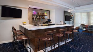 A modern bar area with a flat-screen TV, neon bar sign, and a variety of bottles and glasses. - Courtyard by Marriott Austin Airport (Austin)