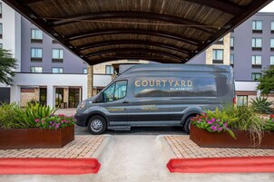 Egy fedett terület alatt parkol egy Courtyard by Marriott márkájú autó, a területhez egy téglából készült járda és virágtartók tartoznak.