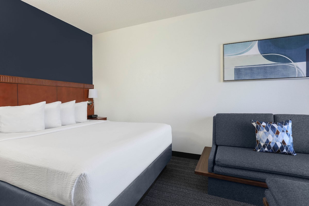 A hotel room with a large bed, a sofa, a nightstand with a lamp, and a wall-mounted abstract painting.