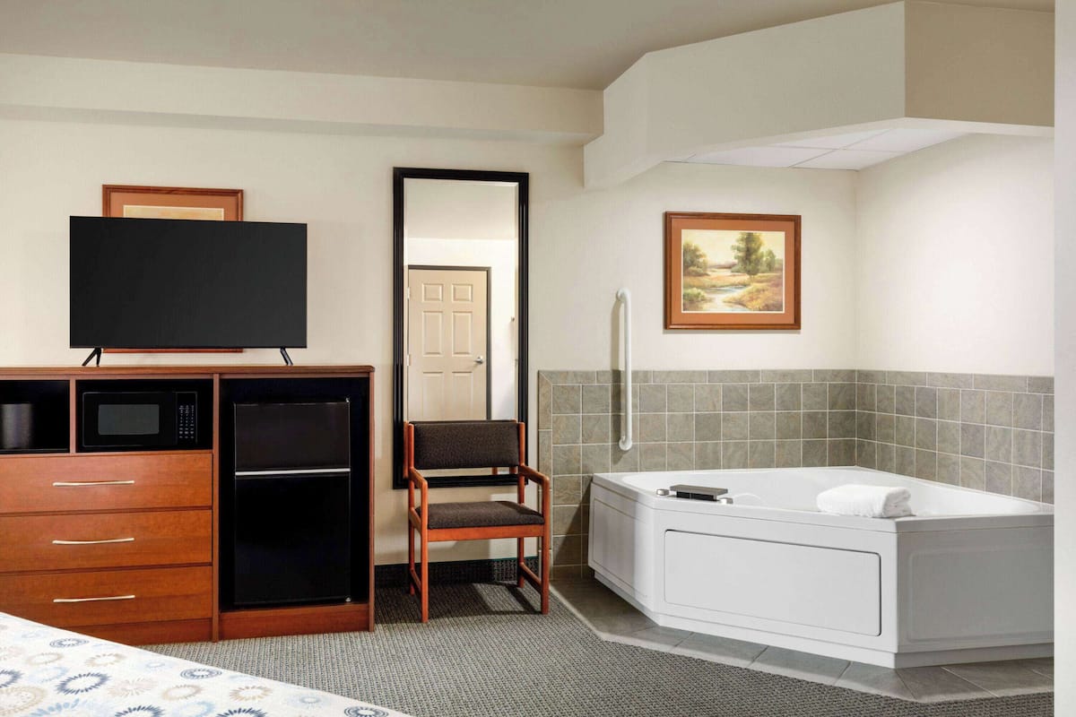 A hotel room with a bed, a TV, a desk with a microwave, a chair, a bathtub, and a painting on the wall.