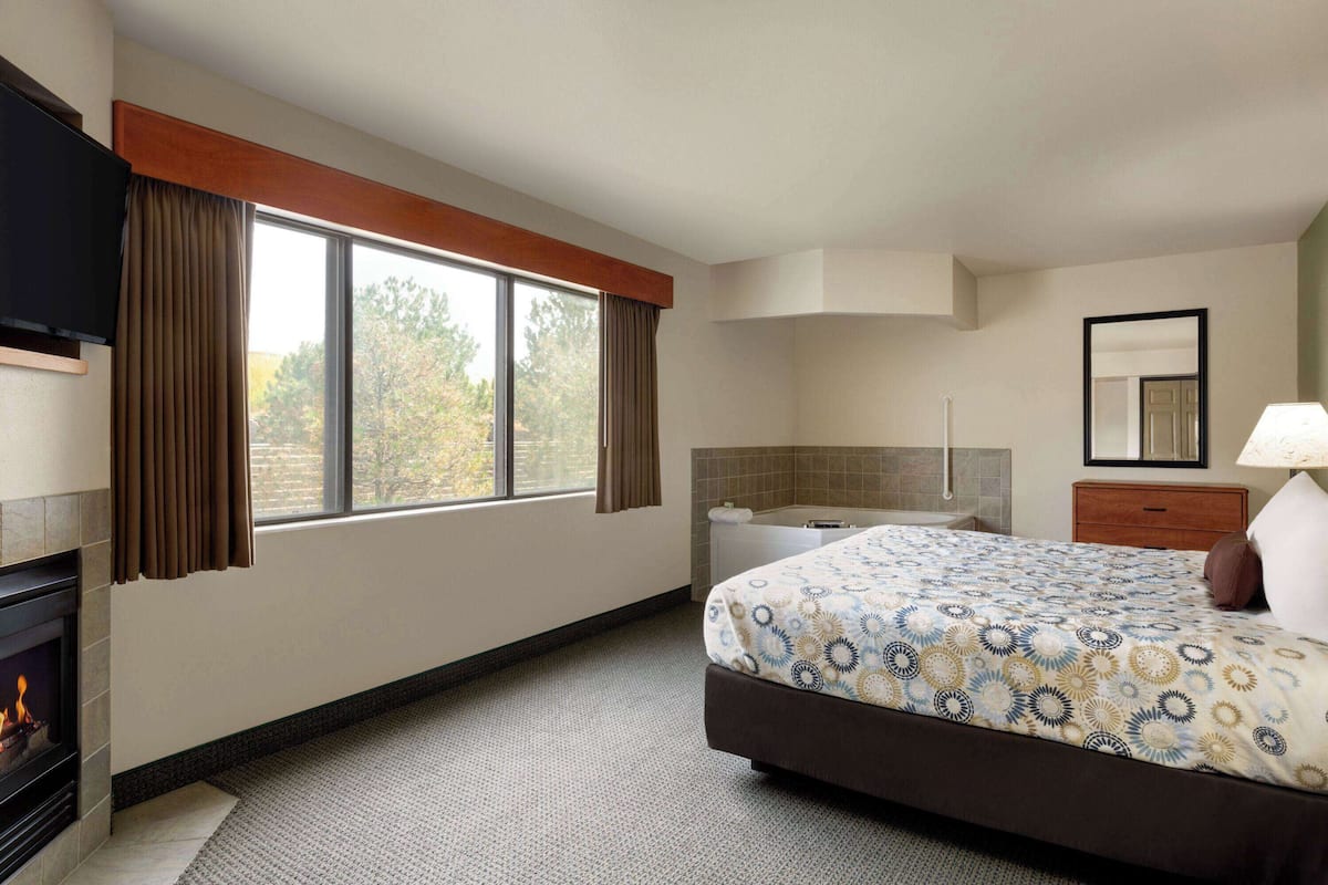 A hotel room with a bed, a TV, a fireplace, a window with curtains, a bathroom, and a mirror.