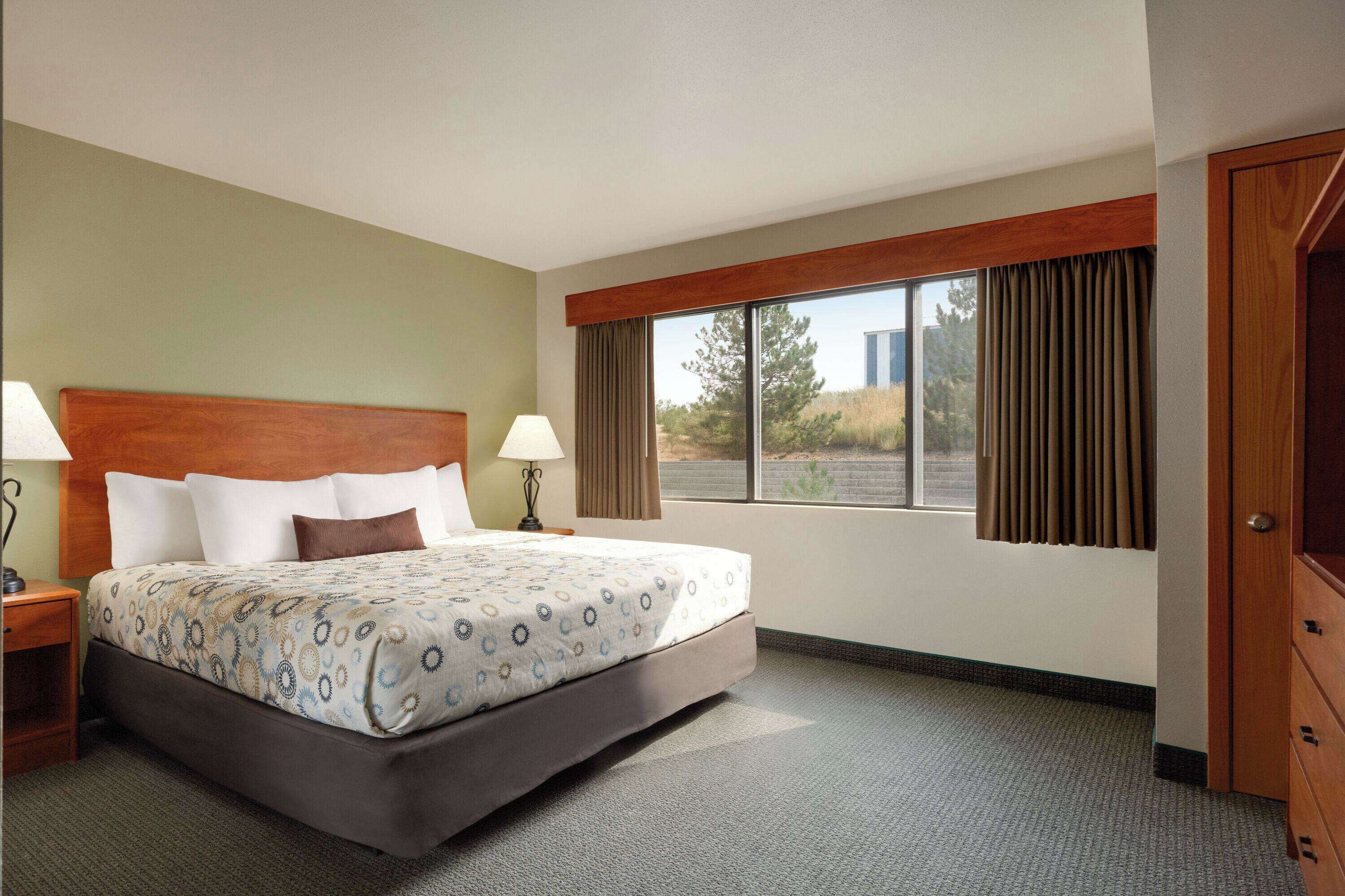 A hotel room with a large bed, two bedside tables with lamps, a window with curtains, and a wooden door.