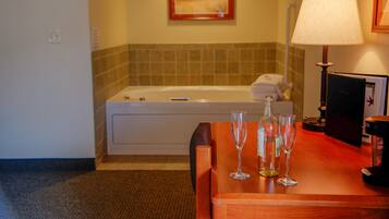A hotel room with a wooden desk, two wine glasses, a bottle, a lamp, a bathtub, and a painting on the wall.